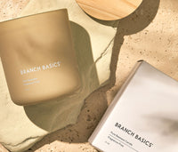 A Branch Basics 100% Beeswax Candle,  laying on its side with light refracting through the glass jar, beside a wooden lid and original packaging, displayed on a neutral stone background. - Thumbnail View 3
