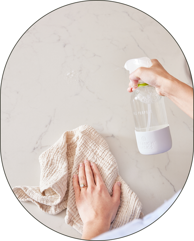 A person using an All-Purpose bottle of Branch Basics Concentrate in their right hand to spray a white marble surface; they are using a lint-free rag in their opposite hand to wipe the surface.
